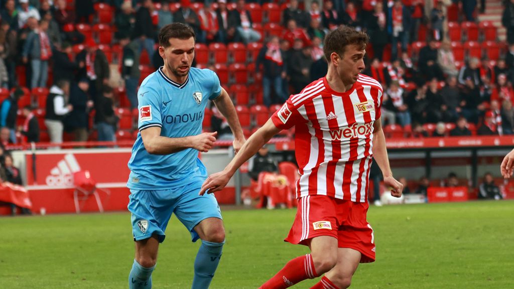 Two soccer players competing during a game, one wearing blue, the other in red and white. Background with fans.