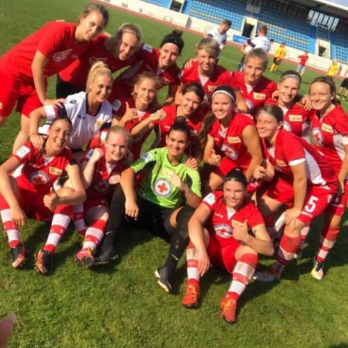 Eine Gruppe von Frauen im roten Fußballoutfit posiert lächelnd auf dem Rasen nach einem Spiel.