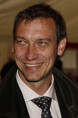 Ein lächelnder Mann mit kurzen, dunklen Haaren trägt ein schwarzes Lederjackett und eine Krawatte.