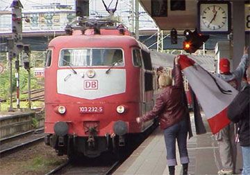 Ein roter Zug der Deutschen Bahn (DB) fährt am Bahnhof ein, während eine Person am Bahnsteig eine Flagge schwenkt.