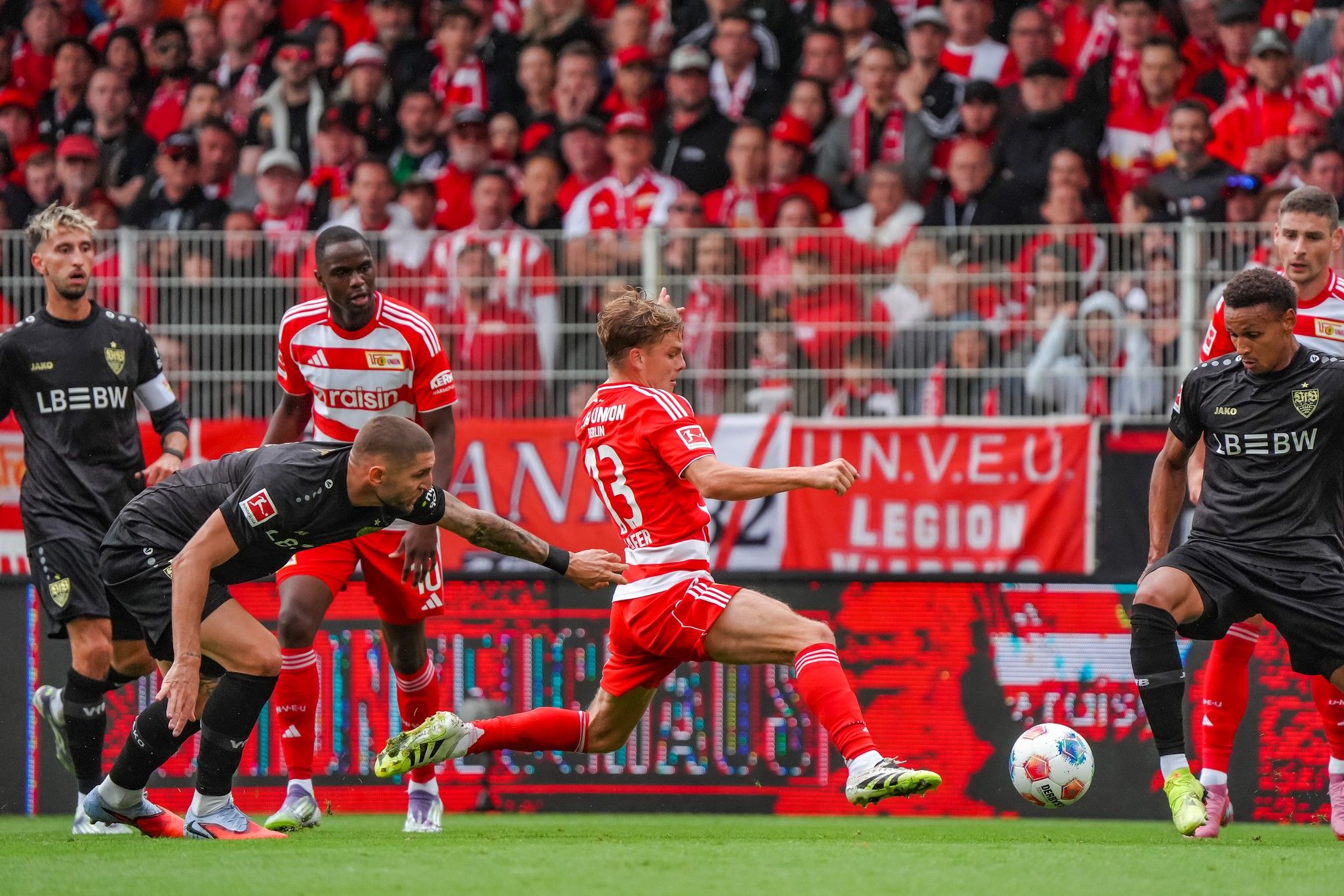 Spielszene aus einem Fußballmatch, zwei Spieler kämpfen um den Ball, Fans im Hintergrund mit Teamflaggen.