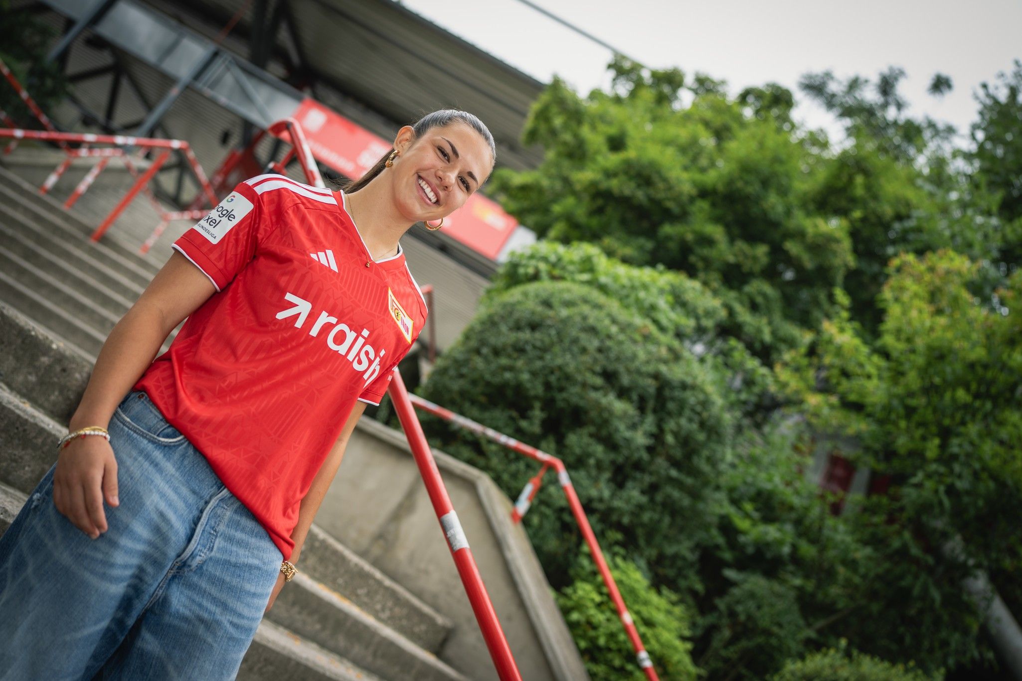 Lächelnde Person in rotem Fußballtrikot steht an einer Treppe mit Grünpflanzen im Hintergrund.