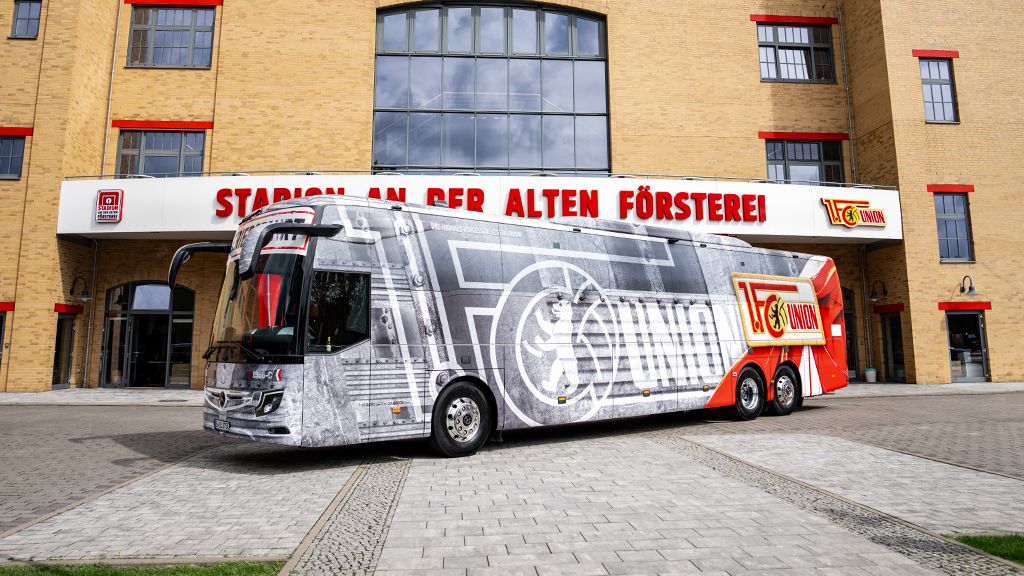 A modern gray bus with a striking design stands in front of a historic red brick building.