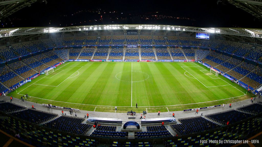 View of an empty, illuminated football stadium with a green field and blue seats.