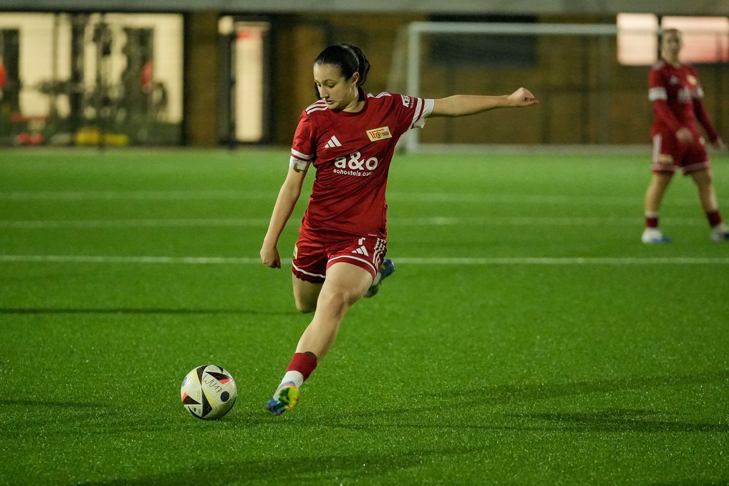 Eine Fußballspielerin in rot tritt einen Ball auf einem Kunstrasenplatz während eines Spiels.