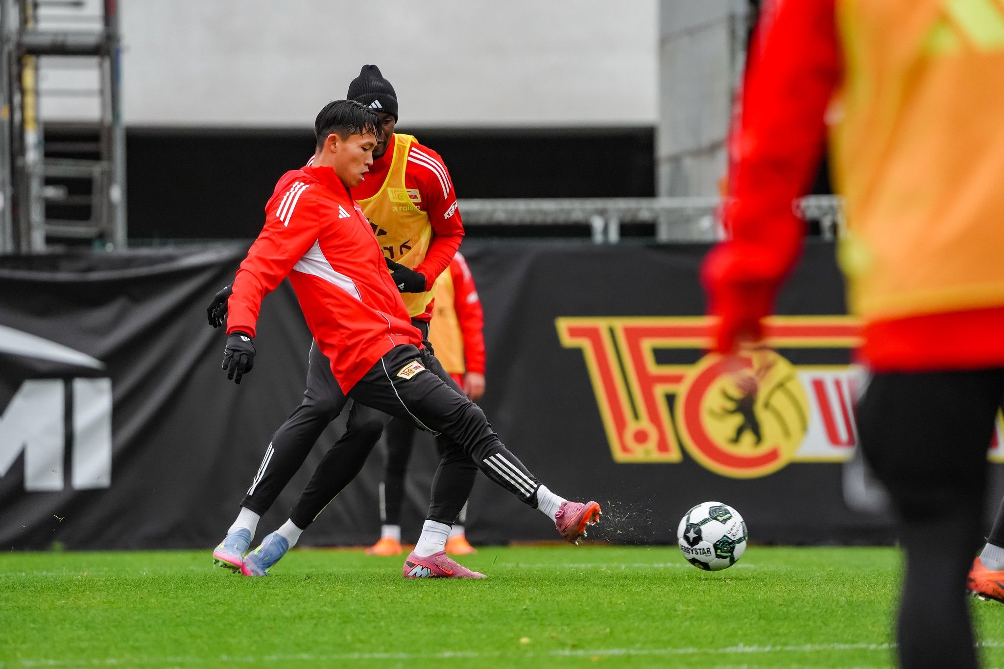 Spieler in roter Trainingskleidung kickt einen Fußball auf einem Trainingsplatz, während ein anderer Spieler ihm nahekommt.