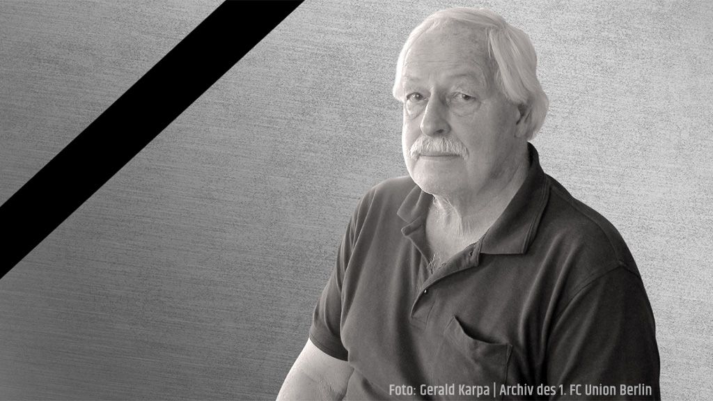 Ein älterer Mann mit grauem Haar und Schnurrbart sitzt in einem dunklen Oberteil vor einem einfarbigen, strukturierten Hintergrund.