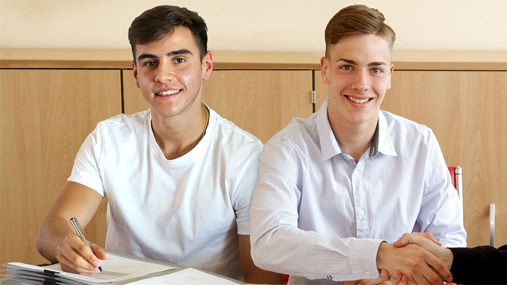 Two young men sit at a table, smiling and shaking hands while signing contracts.