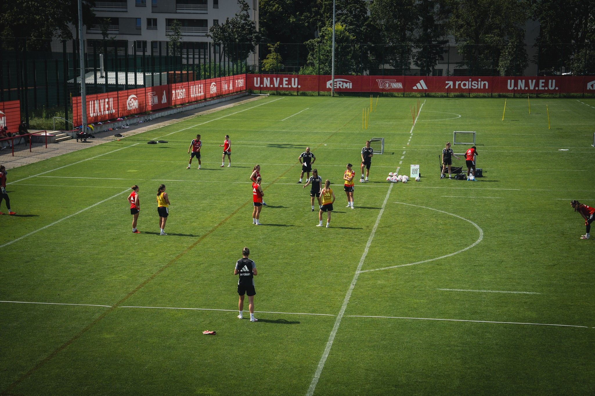 Fußballspieler trainieren auf einem grünen Platz, umgeben von Toren und Trainingsmaterial.