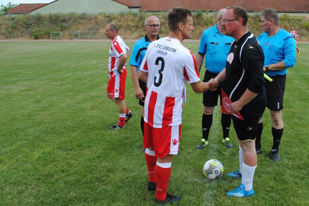 Fußballspieler in rot-weißer Ausstattung schütteln einem Schiedsrichter und einem anderen Spieler die Hand. Rasenfeld im Hintergrund.