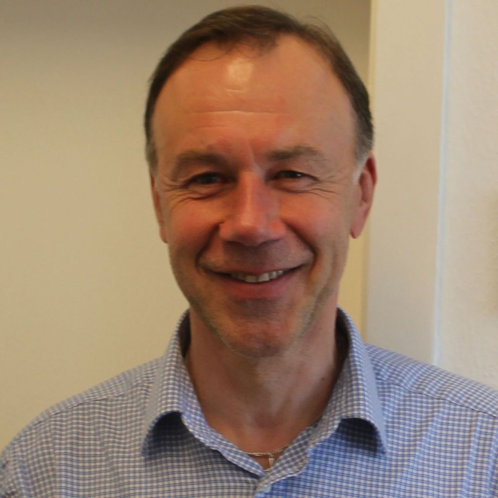 Ein lächelnder Mann trägt ein kariertes Hemd und steht vor einer hellen Wand.