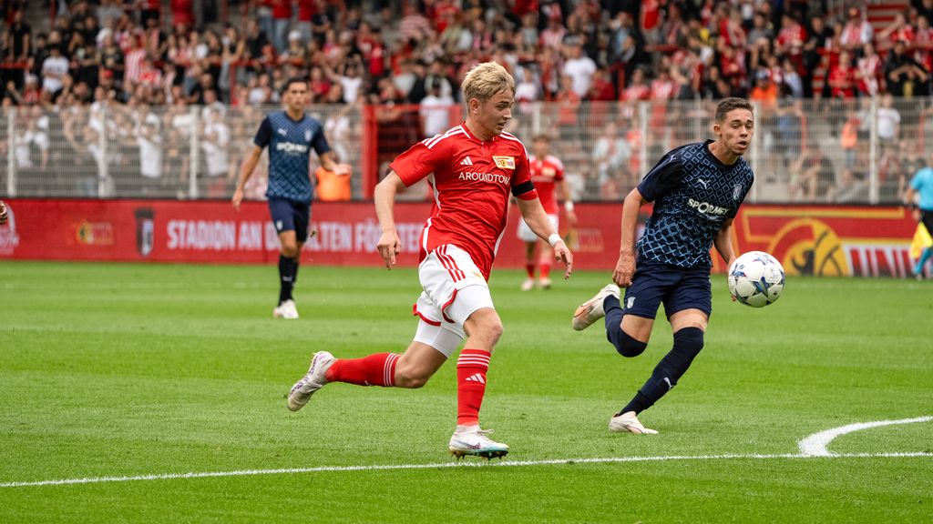 Spielszene im Fußballstadion, ein Spieler in rotem Trikot läuft mit dem Ball, während ein Gegner ihn verfolgt. Fans im Hintergrund.