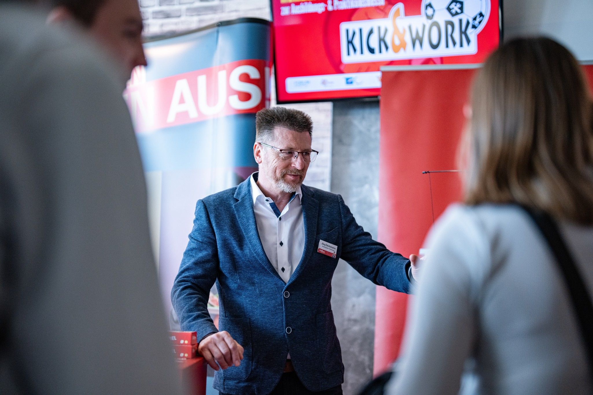 Ein Mann in einem blauen Anzug präsentiert vor einem Bildschirm, während Personen im Vordergrund ihm aufmerksam zuhören.