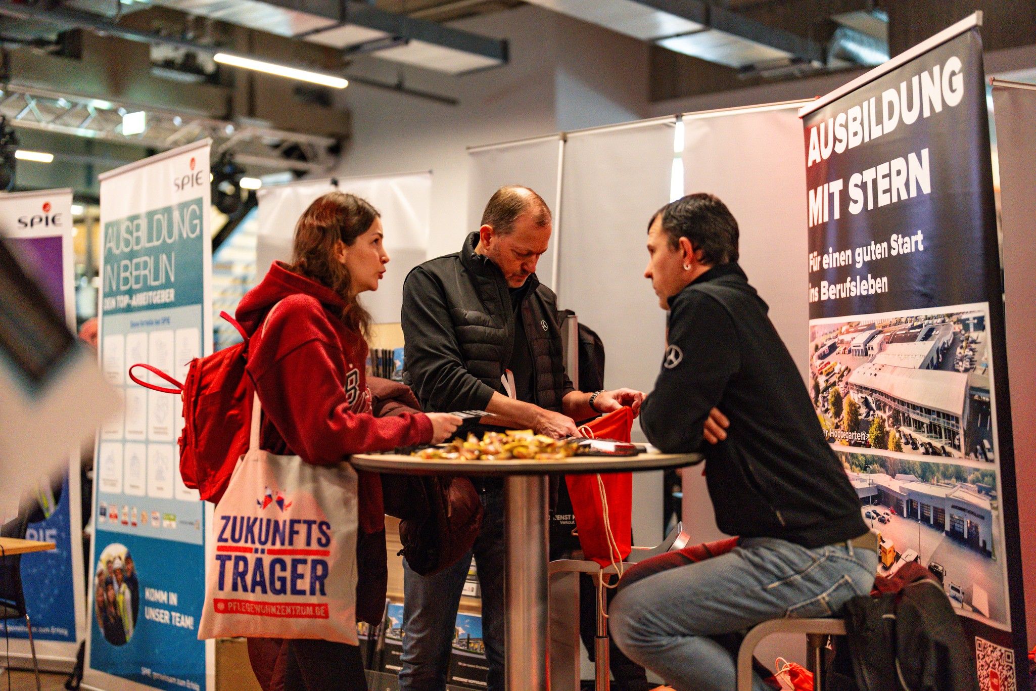 Drei Personen stehen an einem Tisch, während sie Informationen an einem Messestand anschauen. Im Hintergrund sind Banner zu sehen.