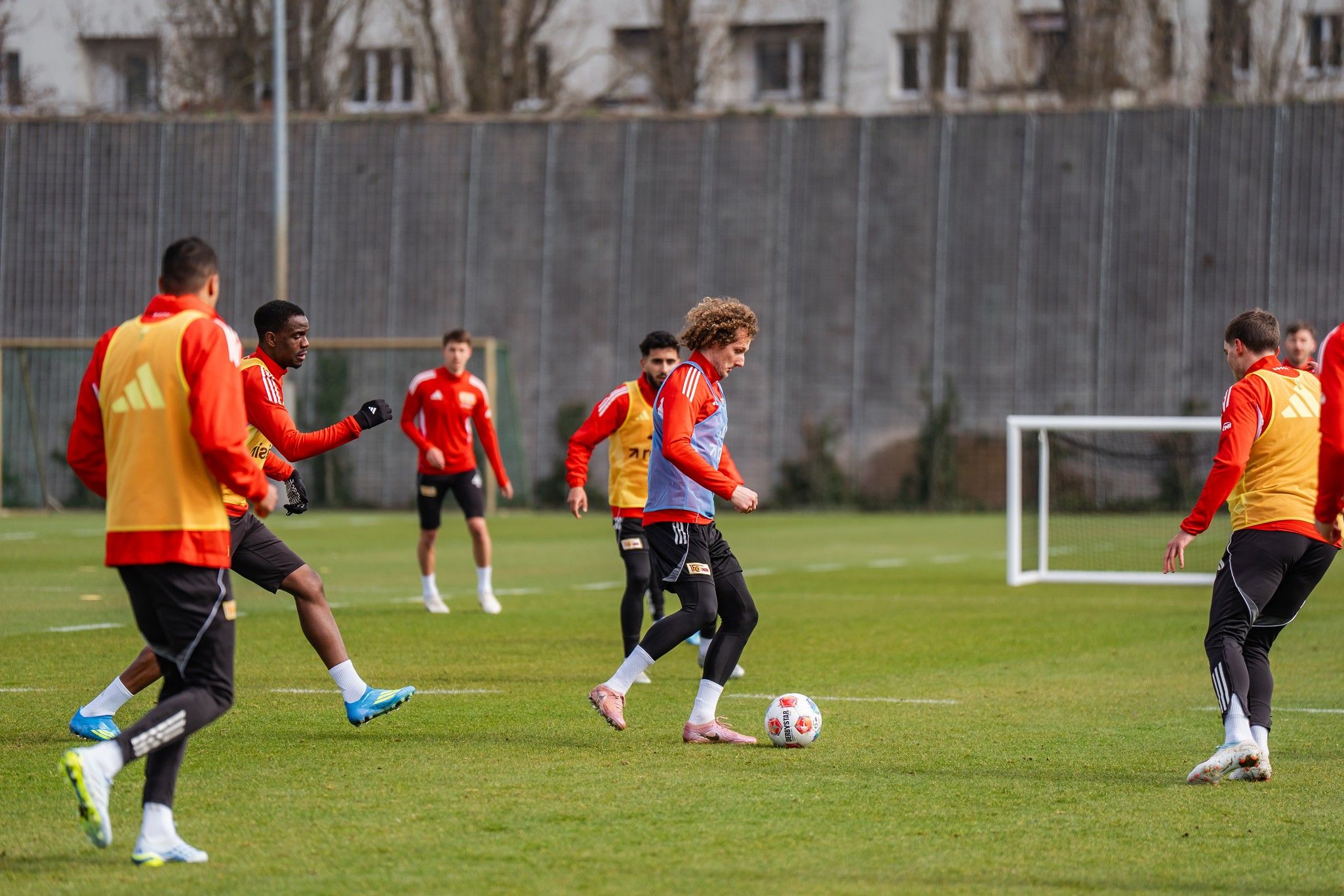 Fußballtraining mit mehreren Spielern auf einem Rasenplatz, einer dribbelt den Ball, während andere in orangefarbener Trainingskleidung agieren.