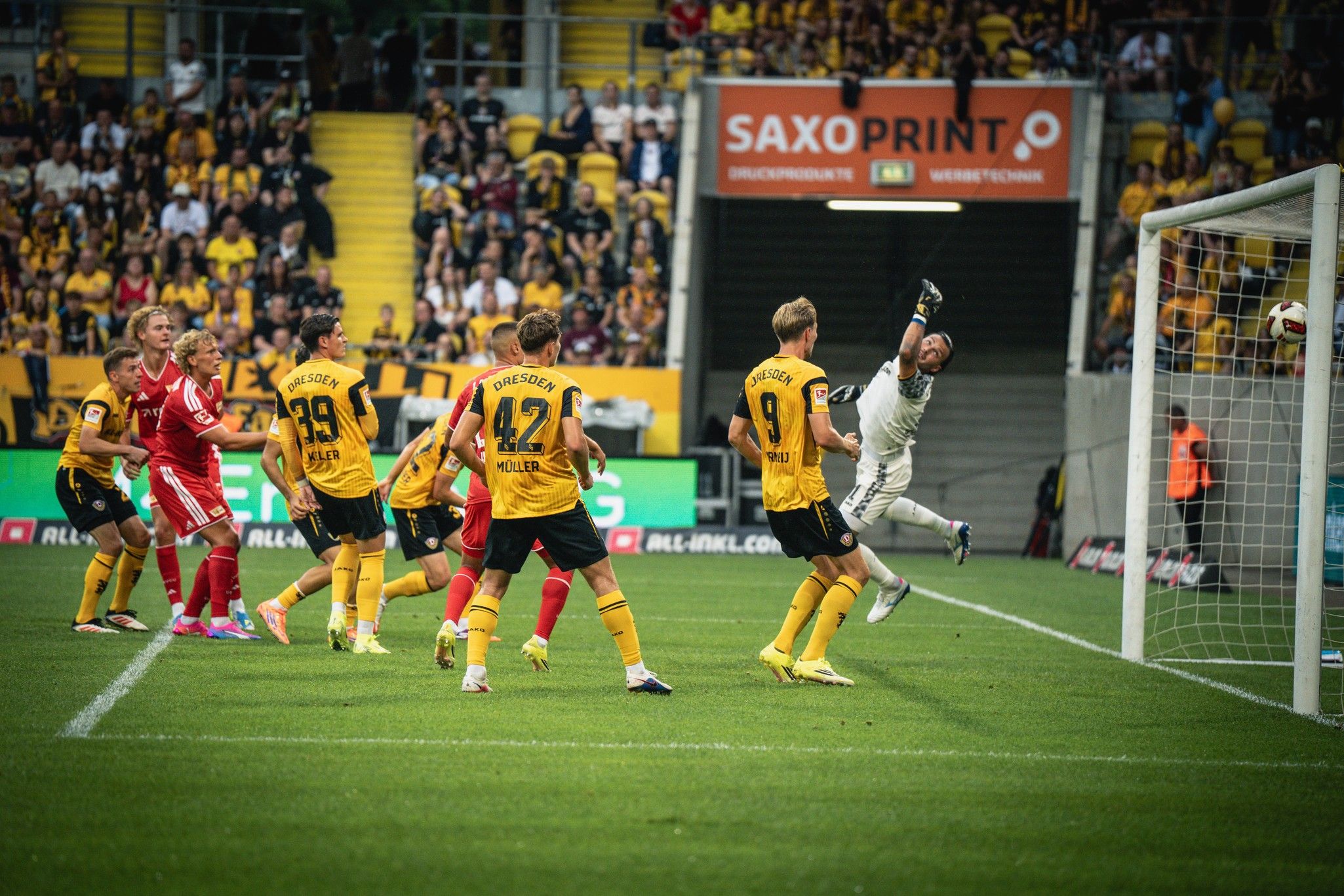 Spielszene mit mehreren Fußballspielern in gelben und schwarzen Trikots, während ein Torhüter einen hohen Schuss abwehrt. Fans im Hintergrund.