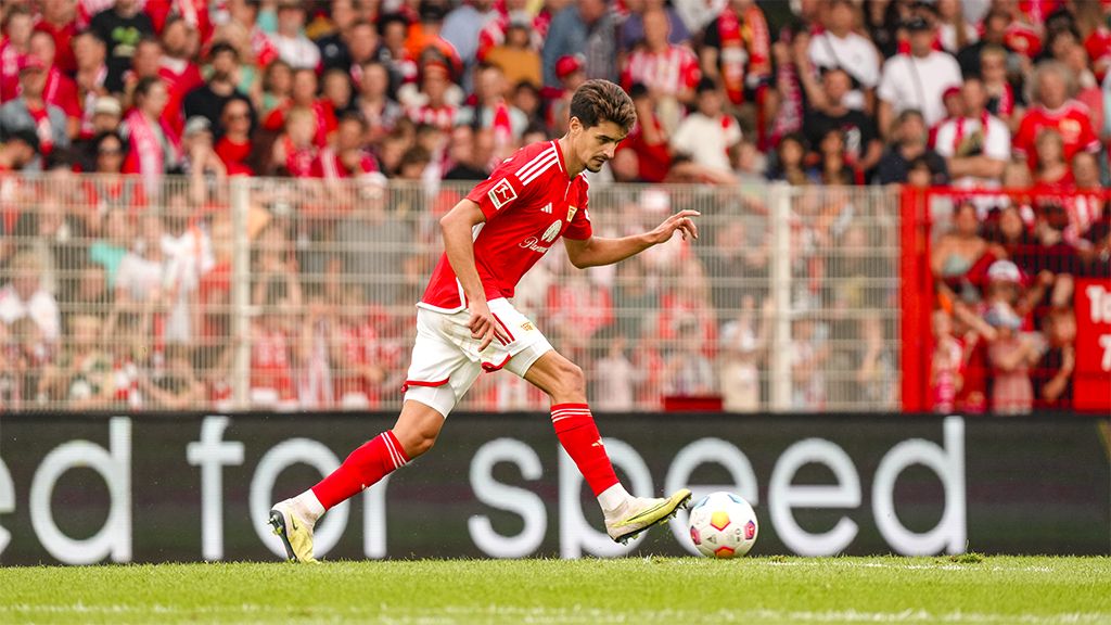 A soccer player in red clothing dribbles the ball on a stadium field, surrounded by cheering fans.