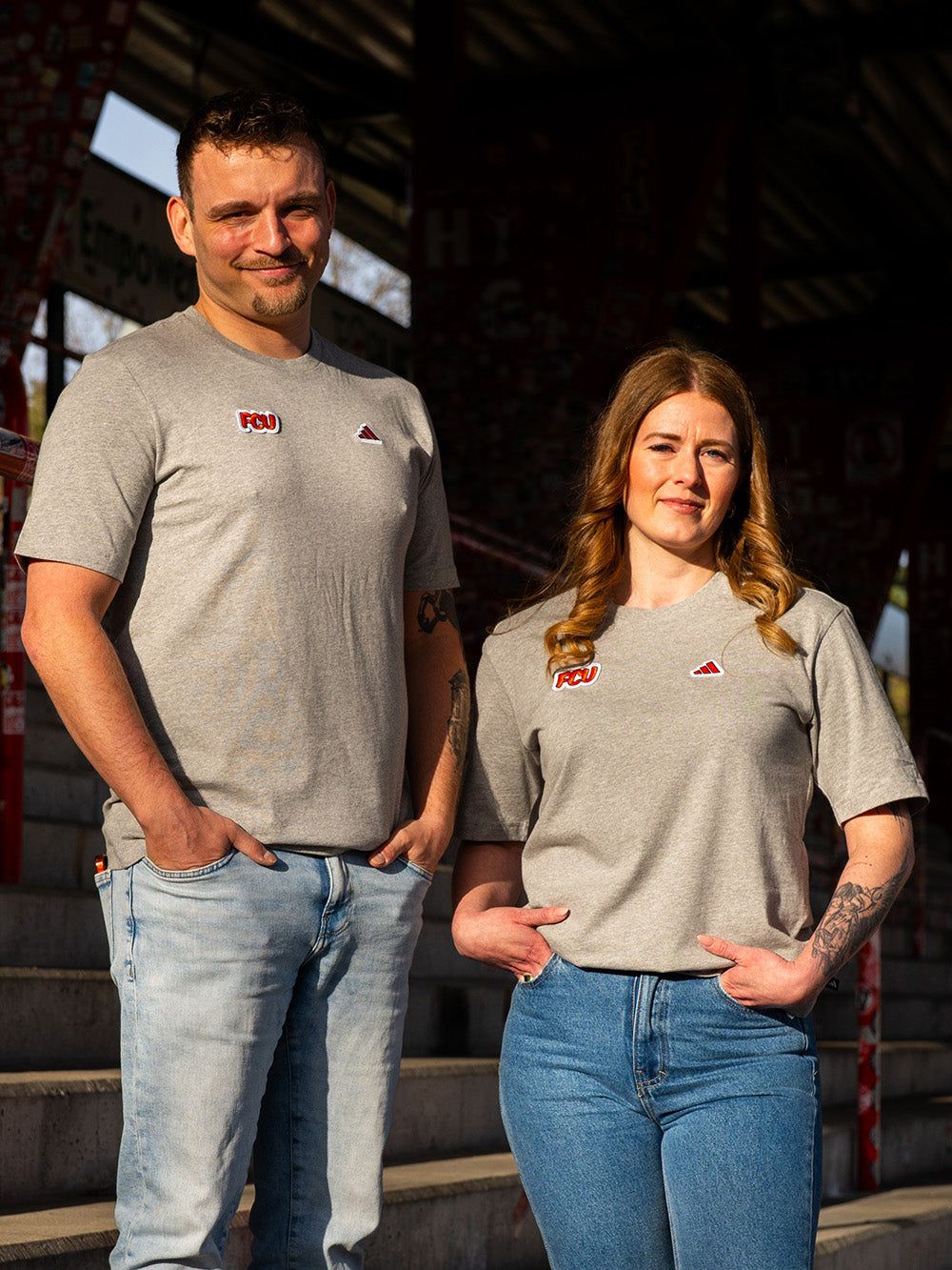 Ein Mann und eine Frau stehen auf einer Treppe, beide tragen graue T-Shirts mit einem Logo und lächeln in die Kamera.