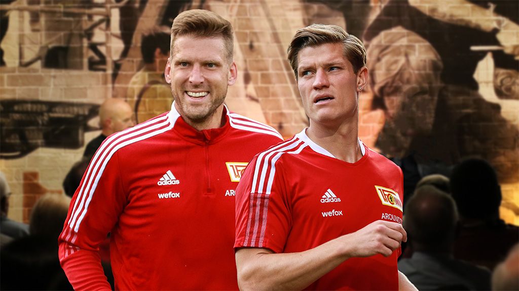 Zwei Fußballspieler in roten Trainingsanzügen mit dem Logo eines Vereins im Hintergrund.