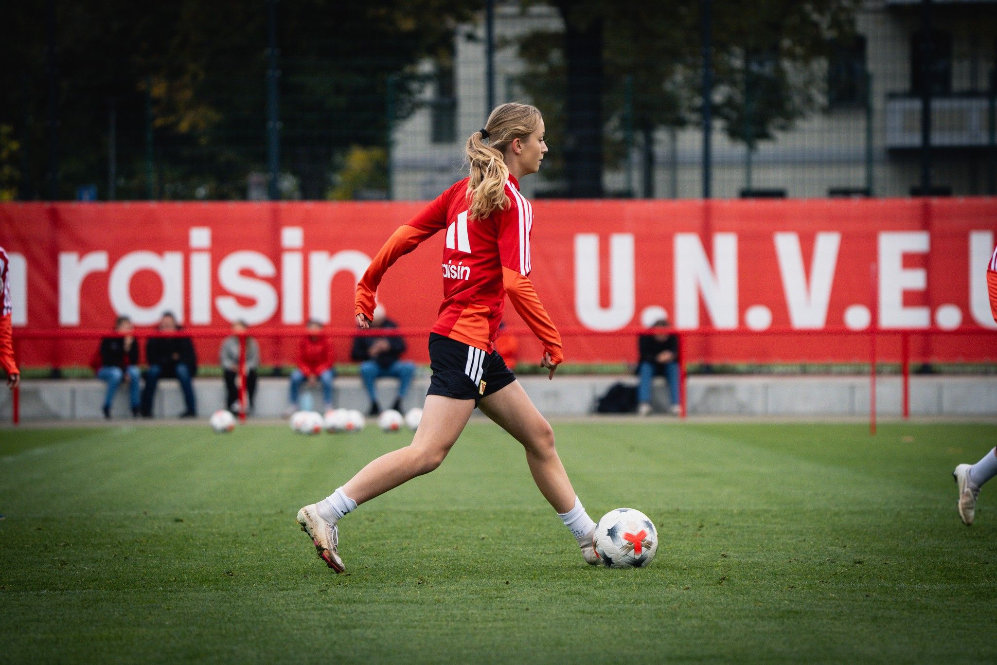 Eine Spielerin in einem roten Trikot läuft mit einem Fußball auf einem Trainingsplatz, im Hintergrund steht ein Banner.