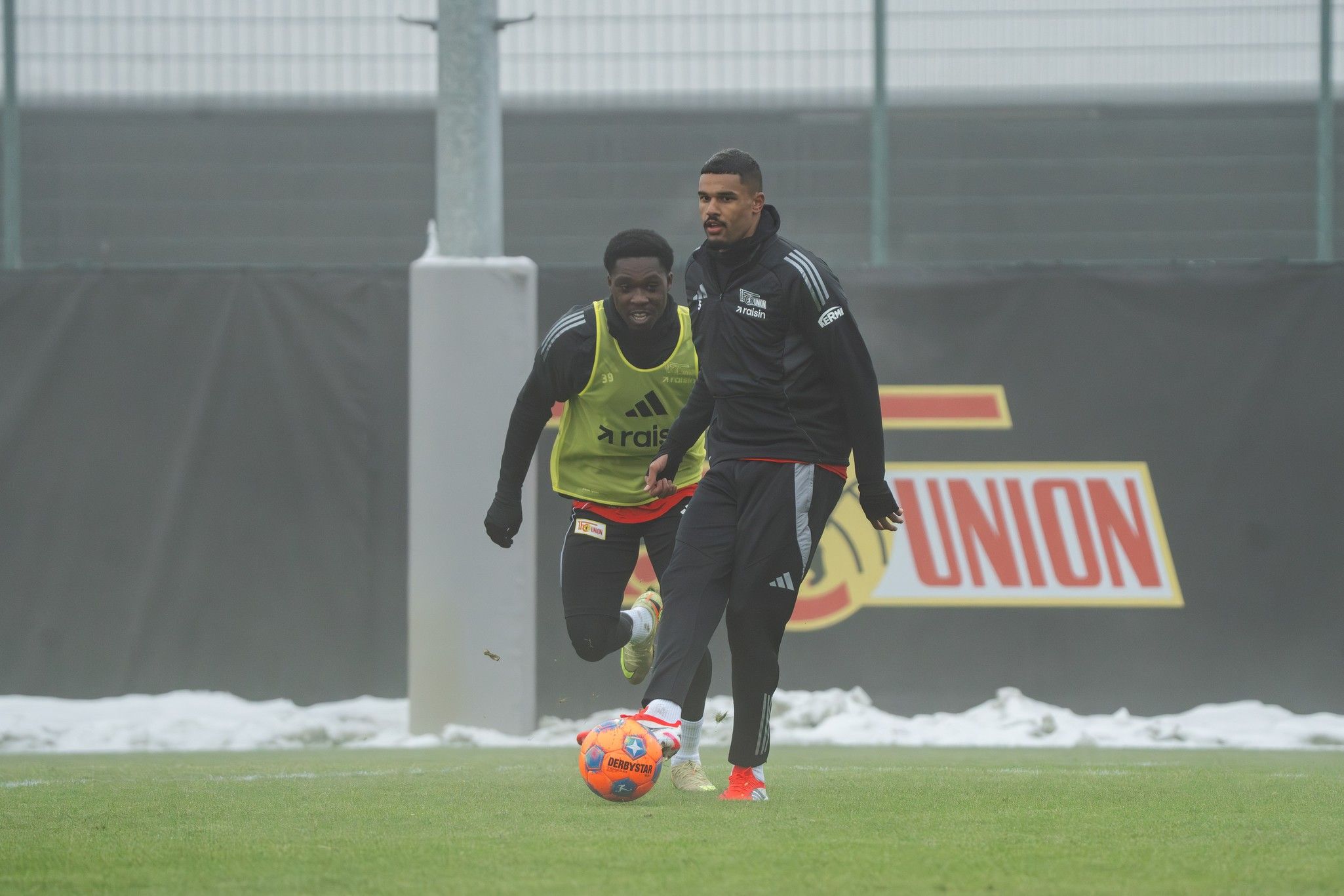 Zwei Fußballspieler trainieren auf einem Platz, einer in einem grünen Trainingsanzug, der andere in schwarzer Sportkleidung.