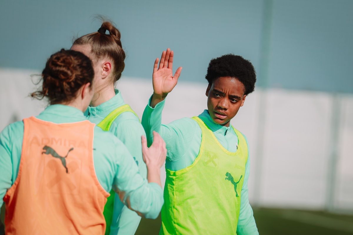 Zwei Fußballspielerinnen in Trainingsbekleidung interagieren auf dem Platz, eine davon hebt die Hand zur Begrüßung oder zum Stoppen.