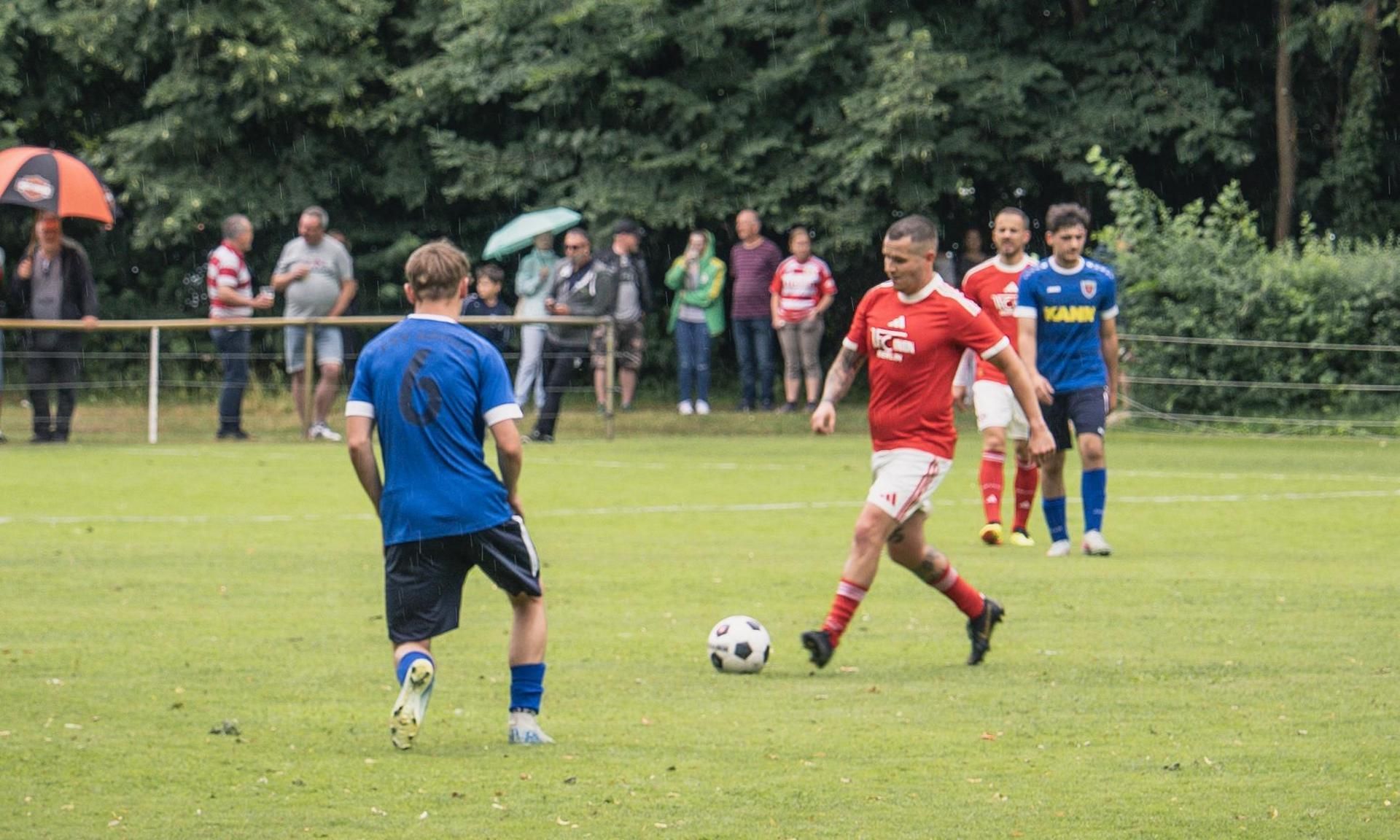 Fussballspiel auf einem grünen Platz mit Spielern in roten und blauen Trikots, Zuschauer im Hintergrund.