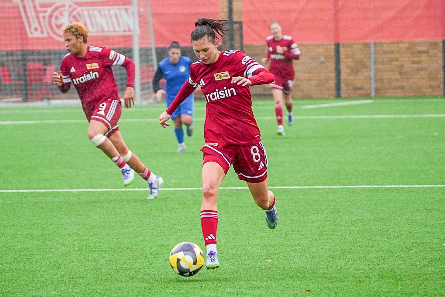 Spielerinnen in roten Trikots kicken auf einem Fußballfeld, während ein Torwart im Hintergrund steht.