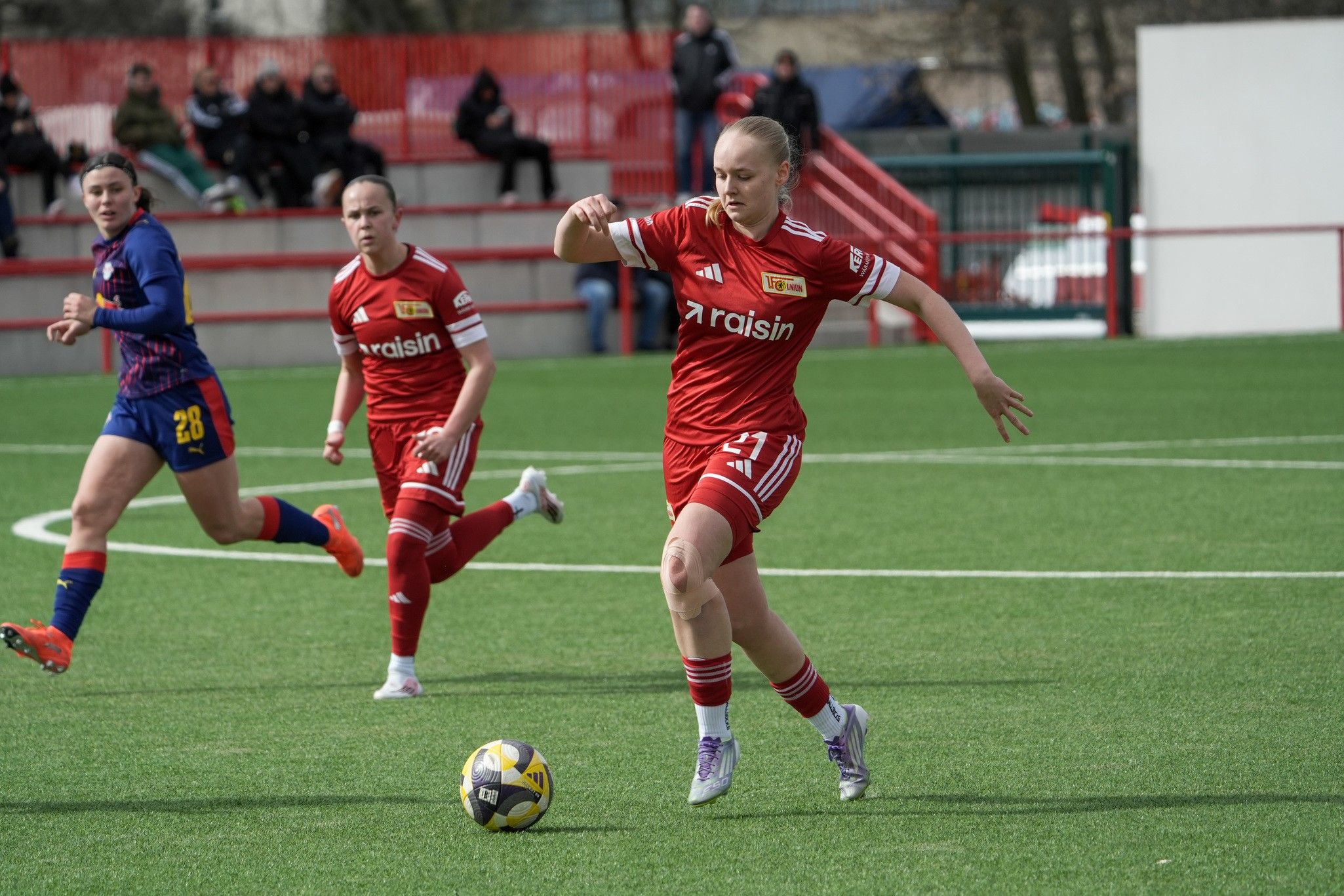 Zwei Fußballspielerinnen in roten Trikots auf einem Kunstrasenfeld, eine Spielerin tritt den Ball.