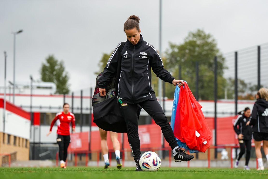 Eine Person in Sportkleidung balanciert einen Fußball und trägt Taschen auf einem Trainingsplatz. Im Hintergrund sind andere Spielerinnen.