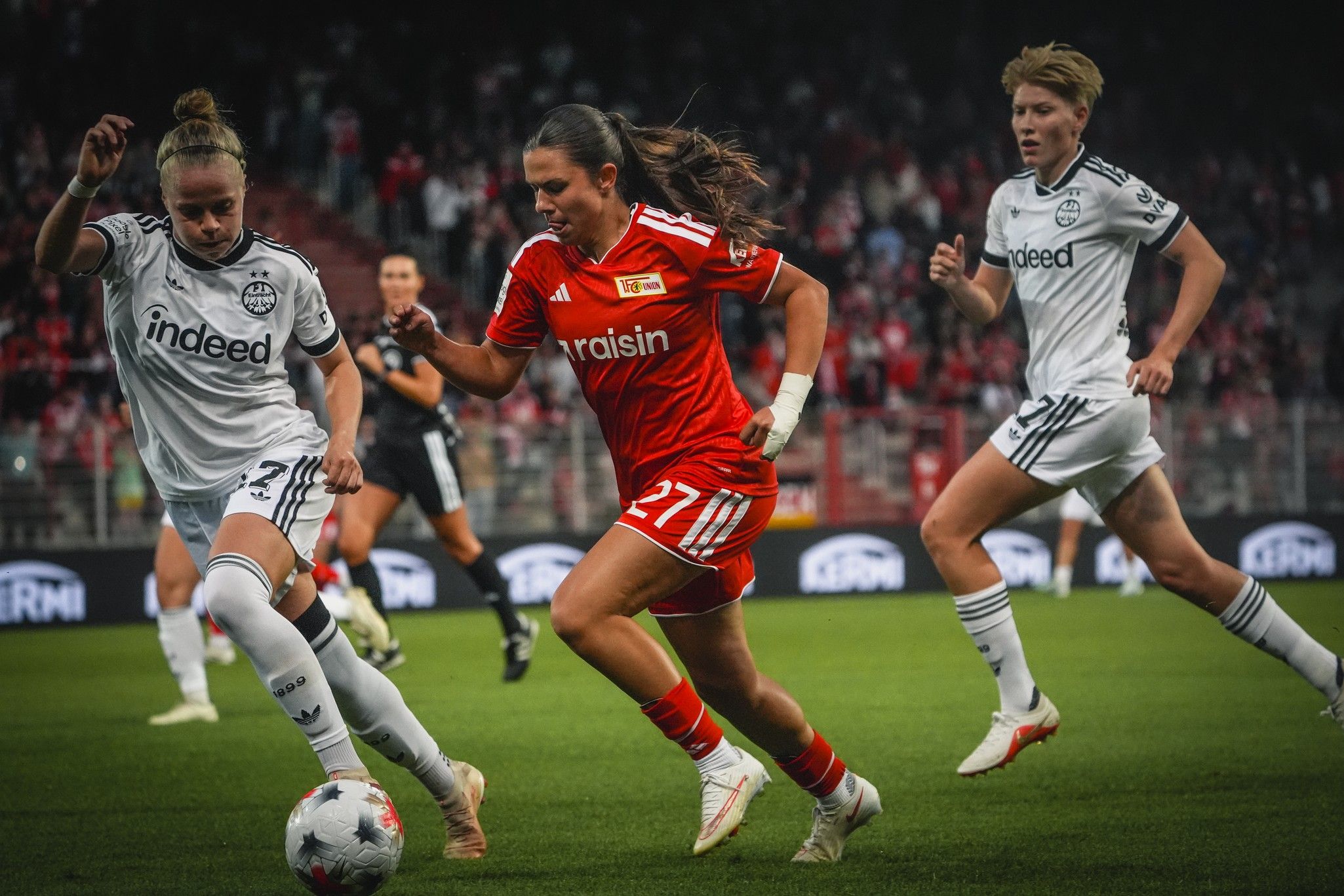 Drei Fußballspielerinnen im Wettlauf auf dem Platz; eine trägt ein rotes Trikot, die anderen zwei weiße Trikots.