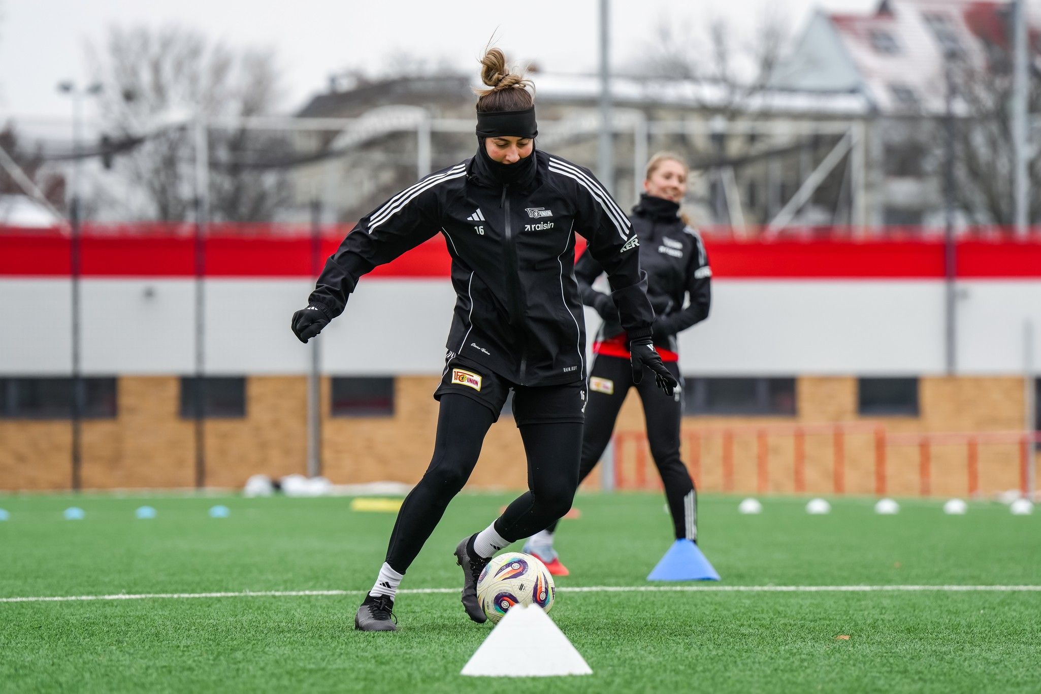 Frauenfußballspielerinnen trainieren auf einem Platz, eine Spielerin dribbelt den Ball um Hütchen. Kühle Wetterbedingungen.