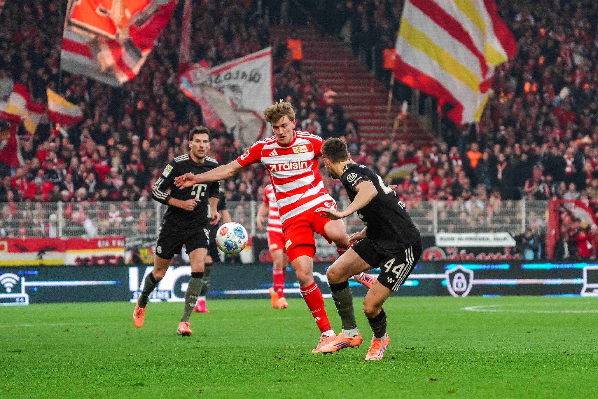 Ein Spieler in rotem Trikot schießt den Ball, während zwei Gegner in schwarz um den Ball kämpfen. Im Hintergrund Fans mit Fahnen.