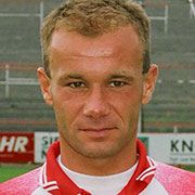 Ein sportlicher Mann mit kurzem, braunem Haar und einem ernsthaften Gesichtsausdruck, trägt ein rotes Sporttrikot.