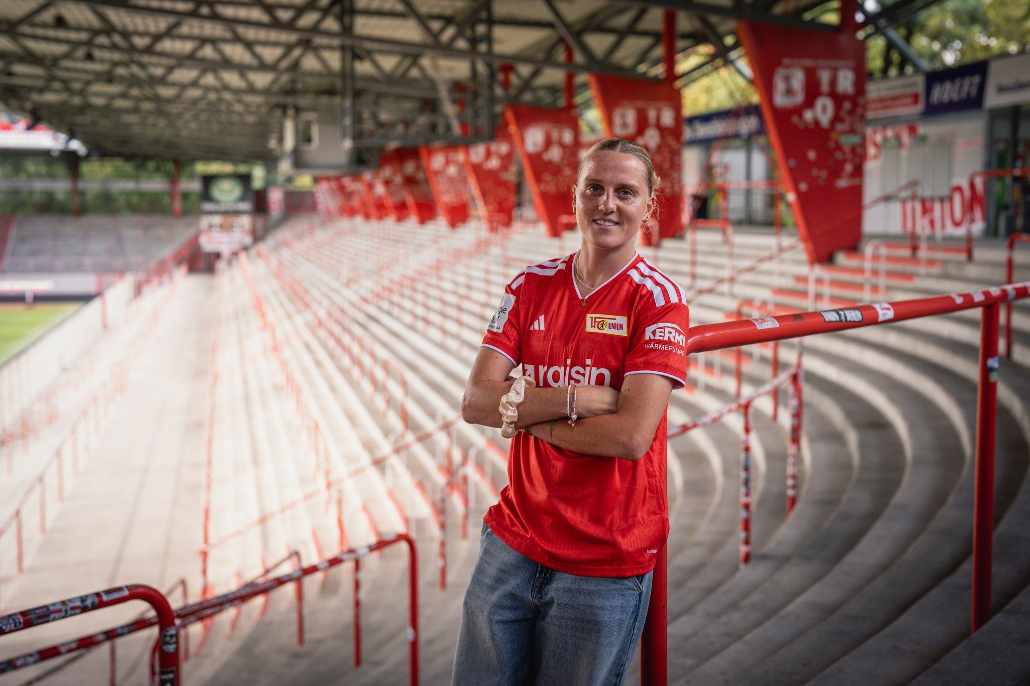 Eine Person in rotem Fußballtrikot steht lächelnd in einem leerstehenden Stadion mit roten Sitzplätzen und Vereinsbanner.