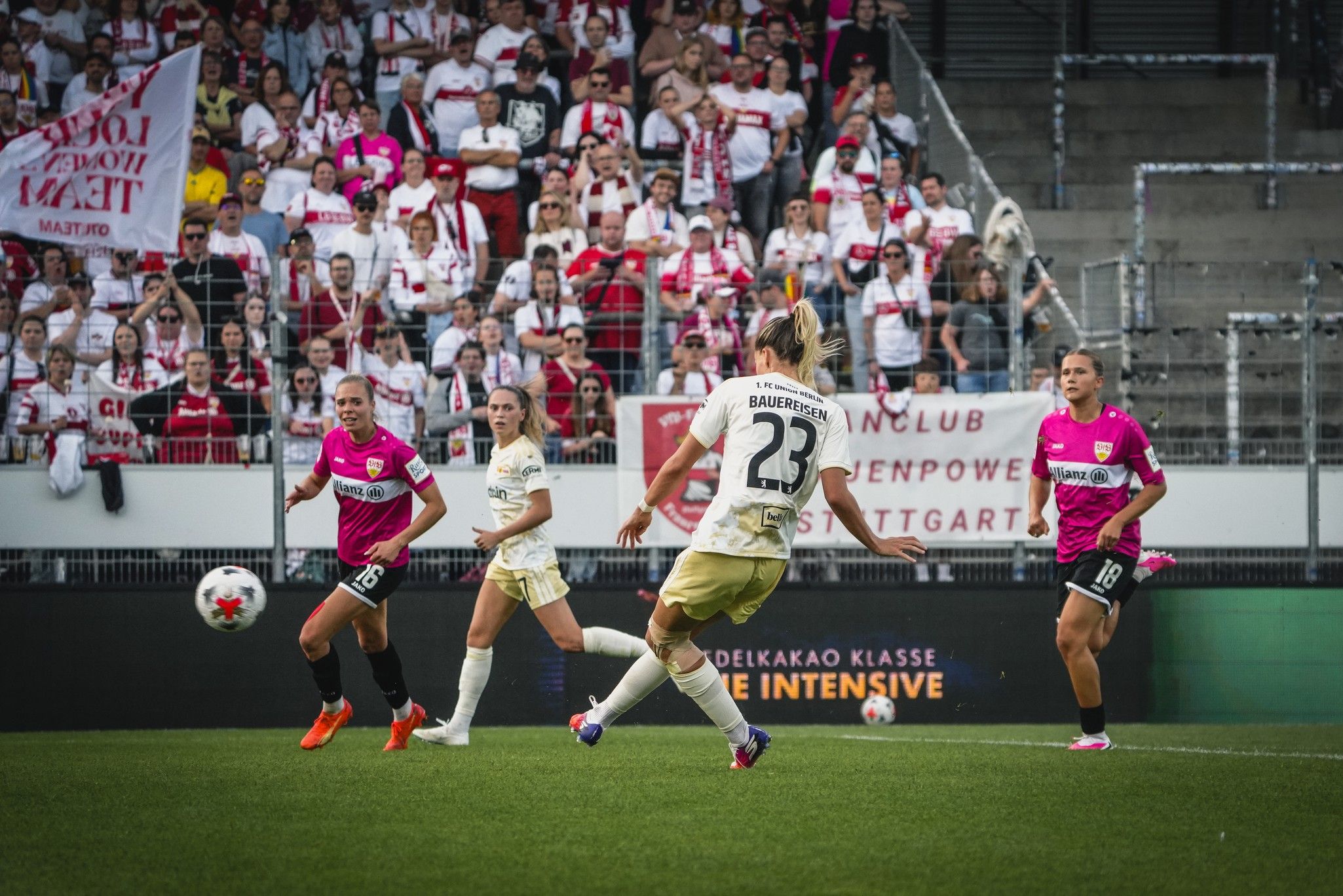 Fußballspiel mit Spielern in weißen und pinken Trikots, Zuschauer im Hintergrund. Spielerin schießt den Ball.