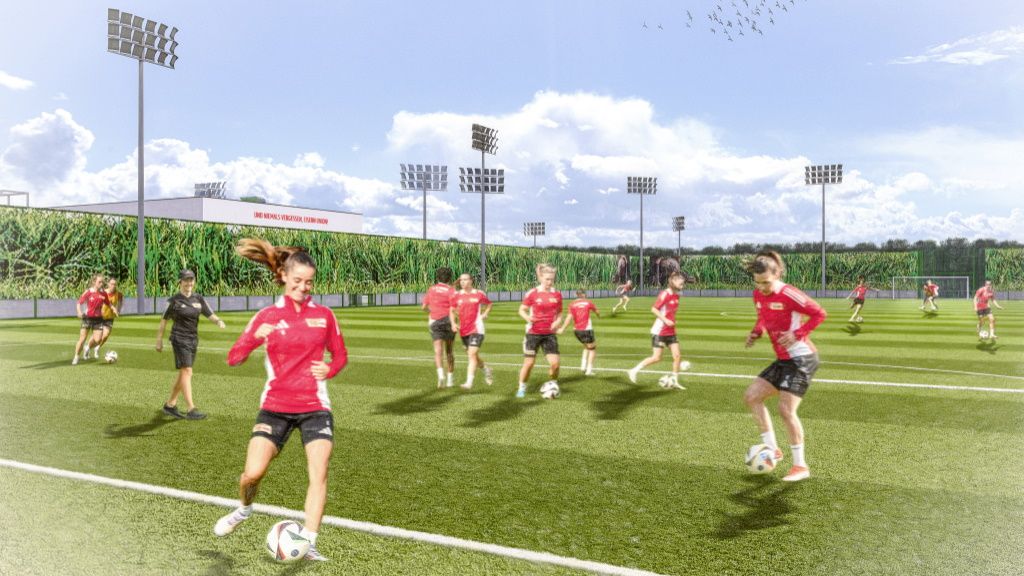 Frauenfußballmannschaft trainiert auf einem Rasenplatz mit Hellblauem Himmel und mehreren Sportbeleuchtungen.