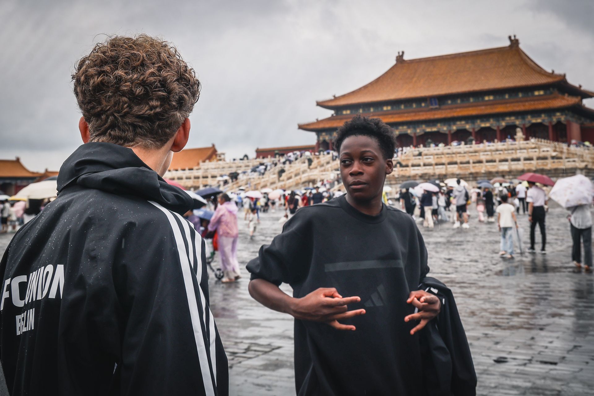 Zwei Personen stehen im Vordergrund vor einer historischen chinesischen Architektur, umgeben von Menschen bei bewölktem Wetter.