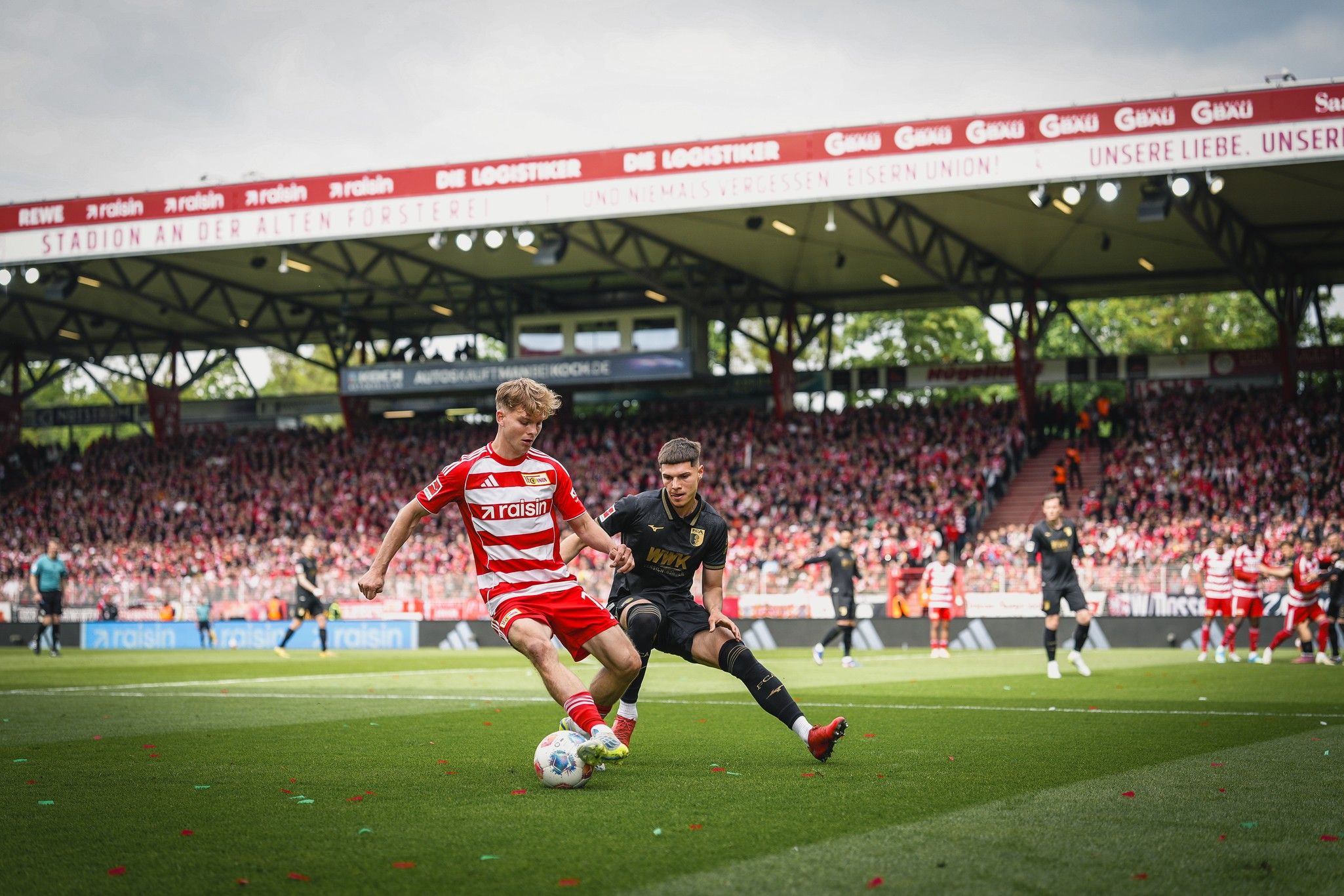 Ein Fußballspieler in rot-weißer Uniform dribbelt den Ball, während ein Gegenspieler in schwarzer Uniform ihn verfolgt. Im Hintergrund eine begeisterte Menge.