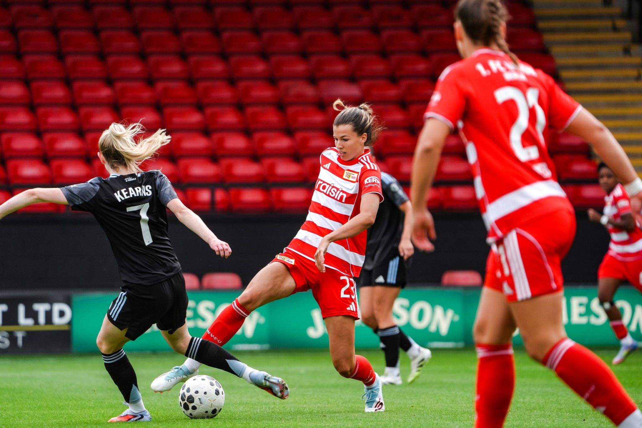 Eine Fußballspielerin in rotem Trikot dribbelt den Ball, während eine Gegnerin in dunklem Trikot versucht, ihn zu erreichen.