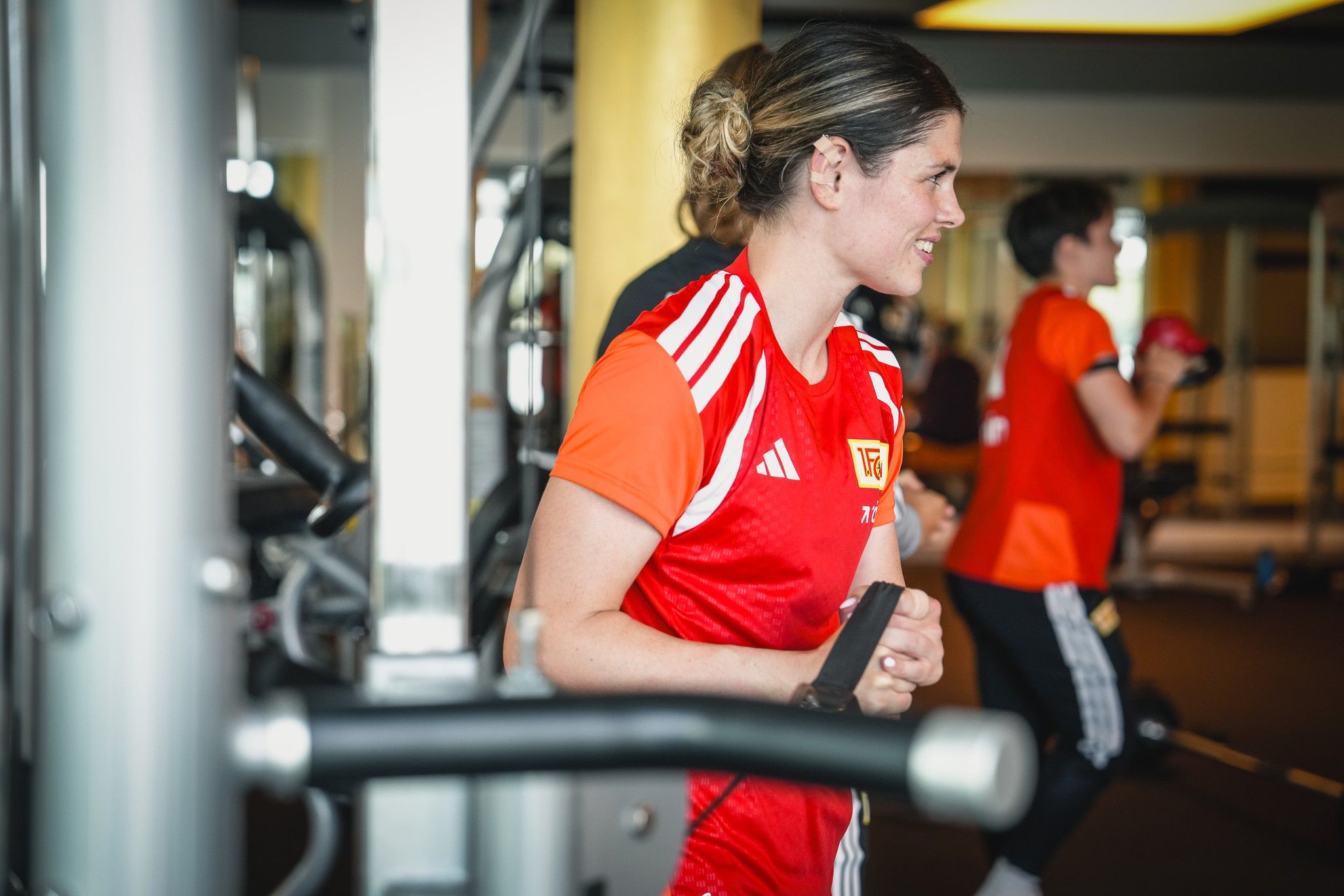 Eine junge Athletin in rotem Sporttrikot trainiert im Fitnessstudio, während eine weitere Person im Hintergrund aktiv ist.