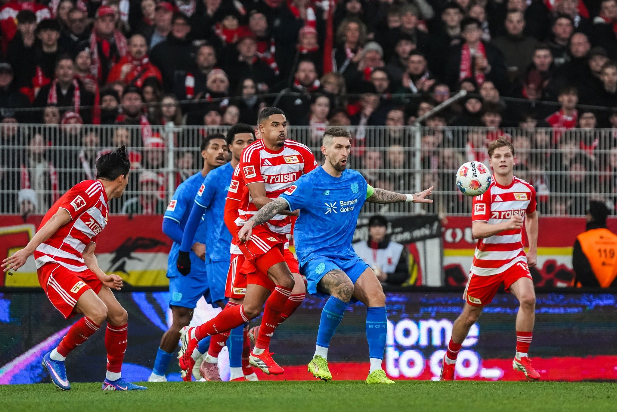 Spielszene im Fußball mit Spielern in roten und blauen Trikots, die um den Ball kämpfen, vor einer begeisterten Menge.