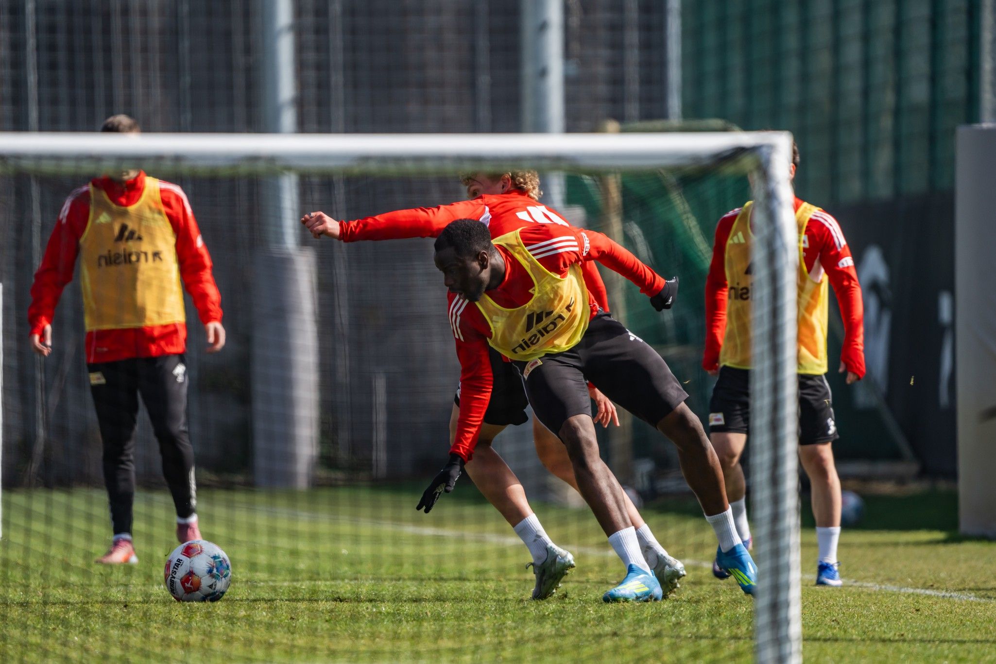 Spieler in gelben Trainingswesten üben auf dem Fußballfeld, während ein Spieler in rot im Hintergrund agiert.