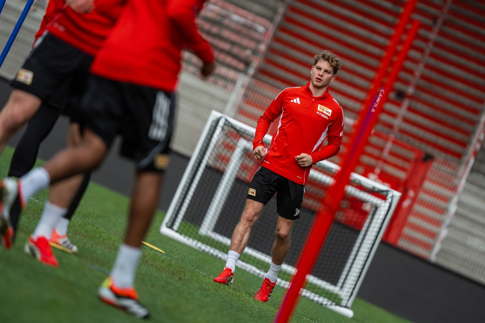 Ein Fußballspieler mit rotem Trikot trainiert auf einem Rasenplatz, umgeben von weiteren Spielern und Fußballtoren.