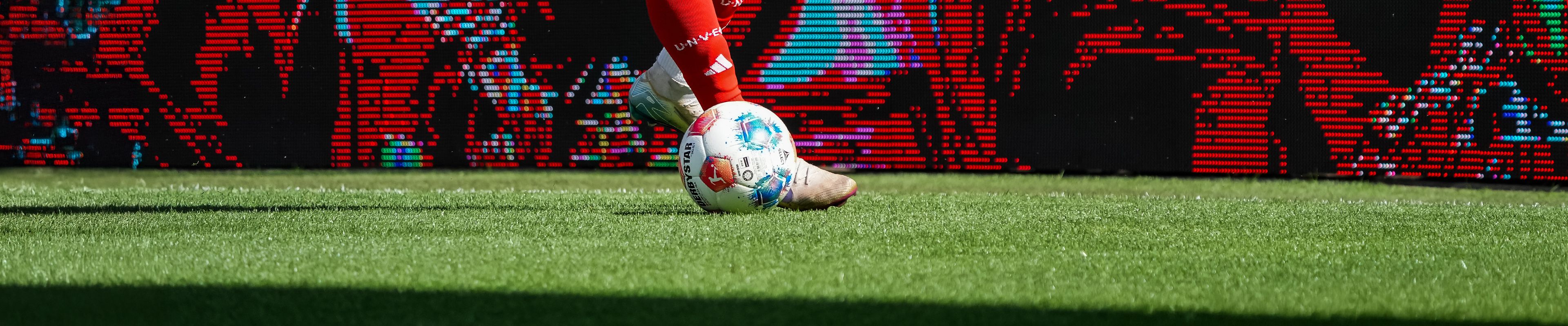 Ein Fußballspieler dribbelt einen bunten Fußball auf einem grünen Rasenfeld, im Hintergrund ist eine digitale Anzeigetafel sichtbar.