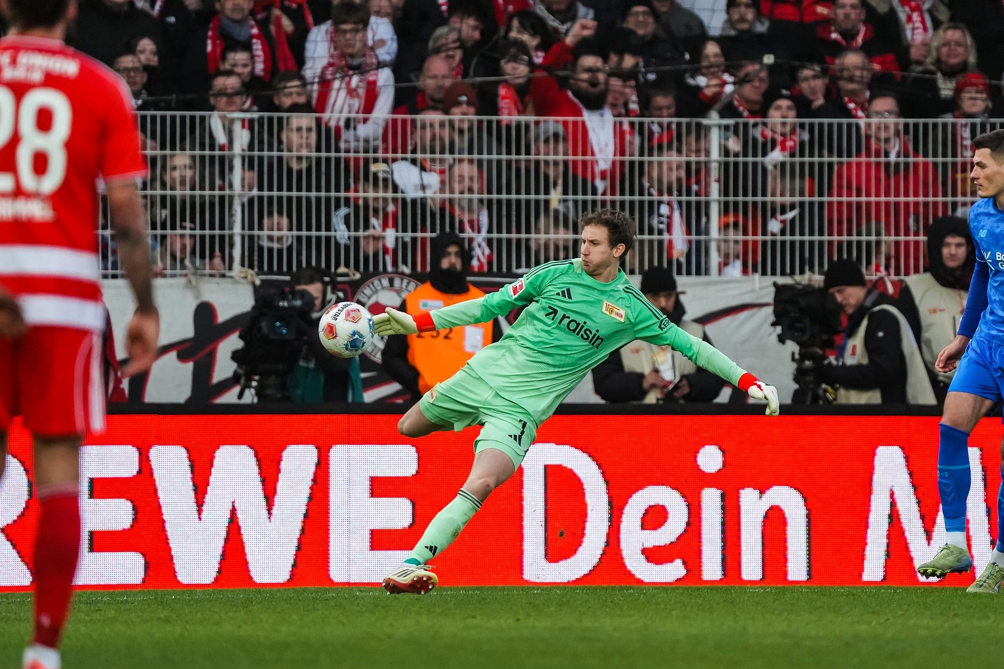 Ein Torwart in grüner Ausrüstung springt, um den Ball während eines Fußballspiels zu fangen, im Hintergrund jubelnde Fans.