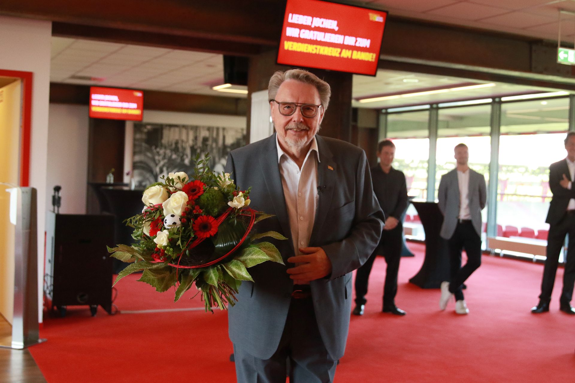 Ein Mann in einem grauen Anzug hält einen Blumenstrauß und steht in einem modernen Raum mit roten Teppich und Bildschirmen im Hintergrund.