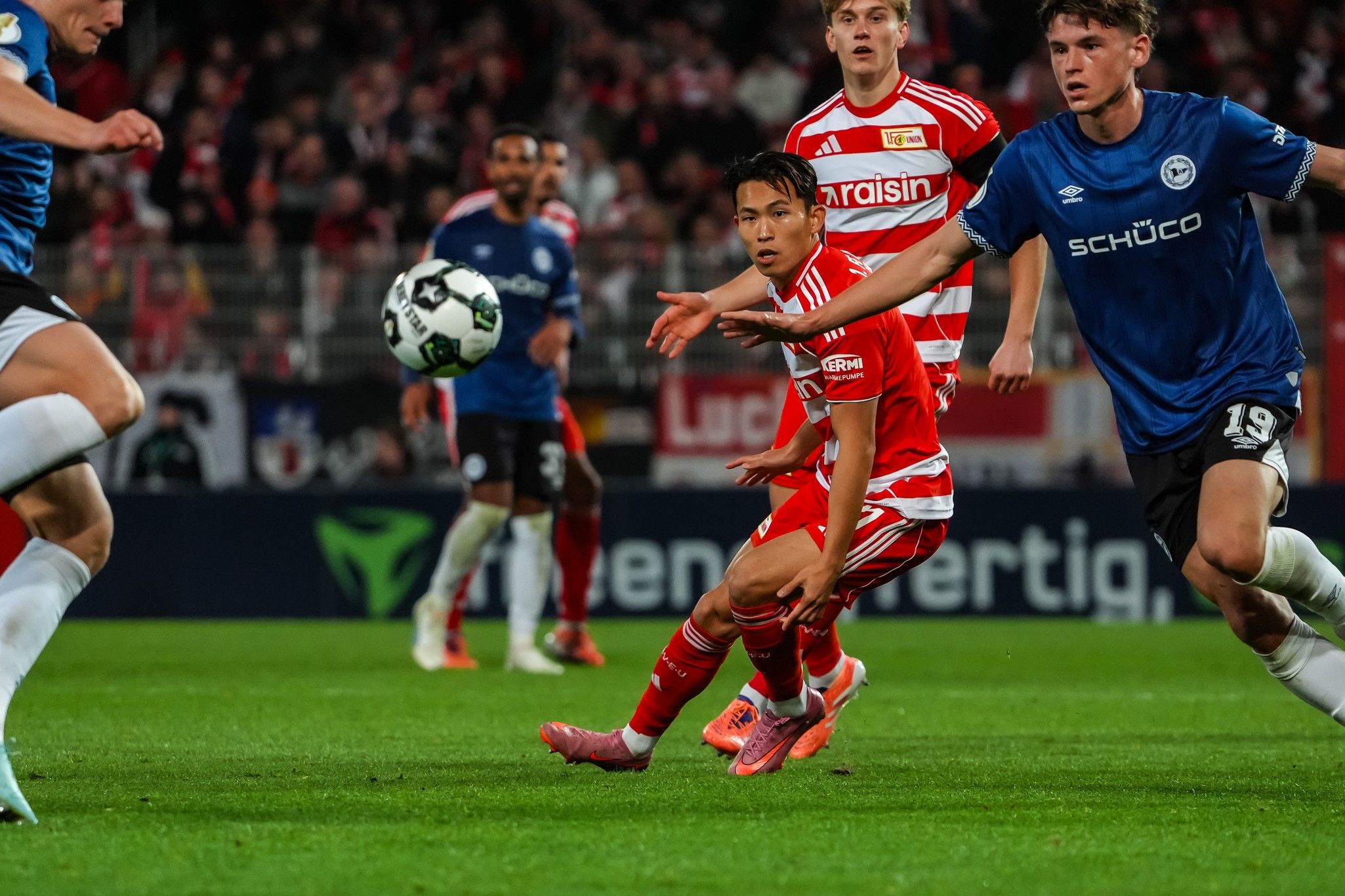 Fußballspiel im Stadion mit Spielern in roten und blauen Trikots, ein Spieler mit dem Ball in Aktion.