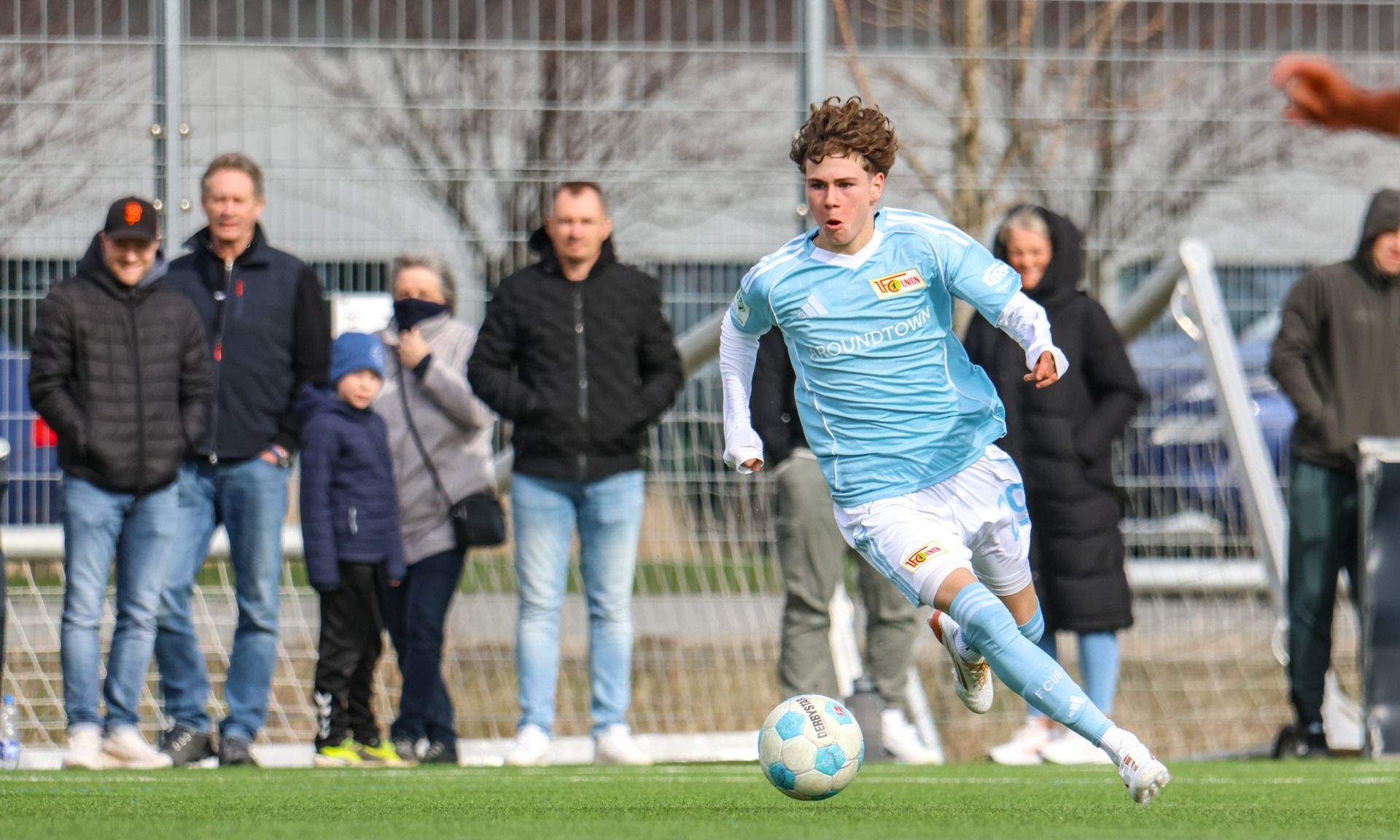 Ein junger Fußballspieler in hellblauer Kleidung sprintet mit dem Ball auf dem Rasen, während Zuschauer im Hintergrund zuschauen.
