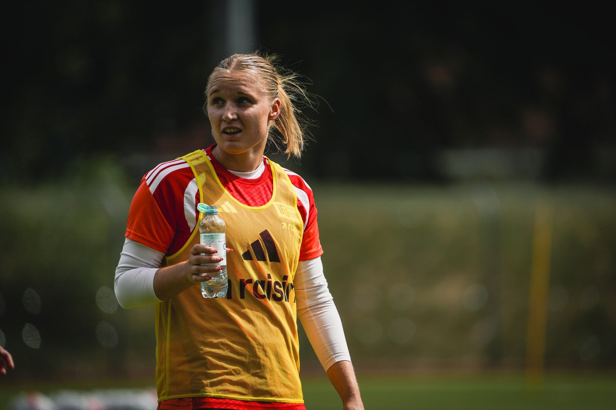 Sportlerin mit Wasserflasche in der Hand, trägt ein gelbes Trainingsoberteil und schaut in die Kamera. Hintergrund mit unscharfer Sportumgebung.